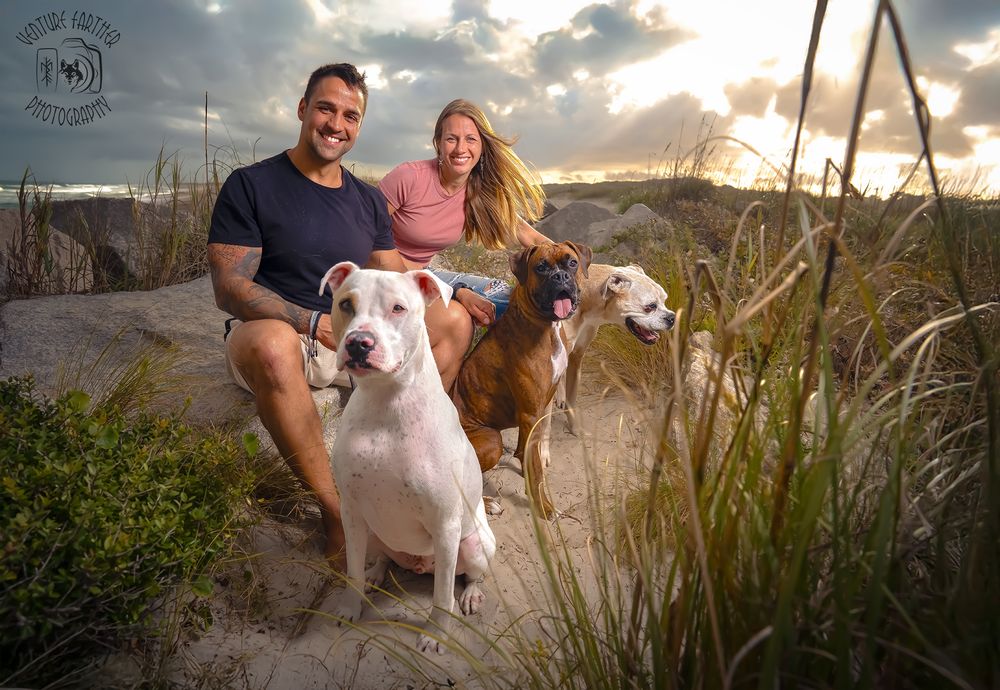 A couple sits with their two dogs on a beach path at sunset with coastal grasses in the foreground.