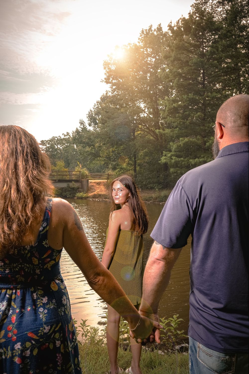 Group of people standing by a lake at sunset with golden light filtering through trees in the background.