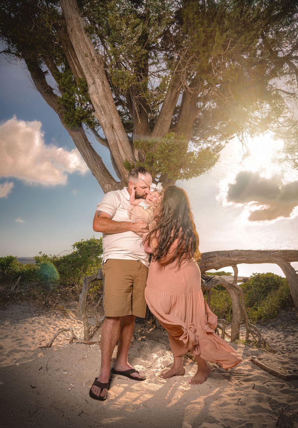 Romantic beach photoshoot with a couple sitting on a tree branch at sunset in matching pink and tan outfits.