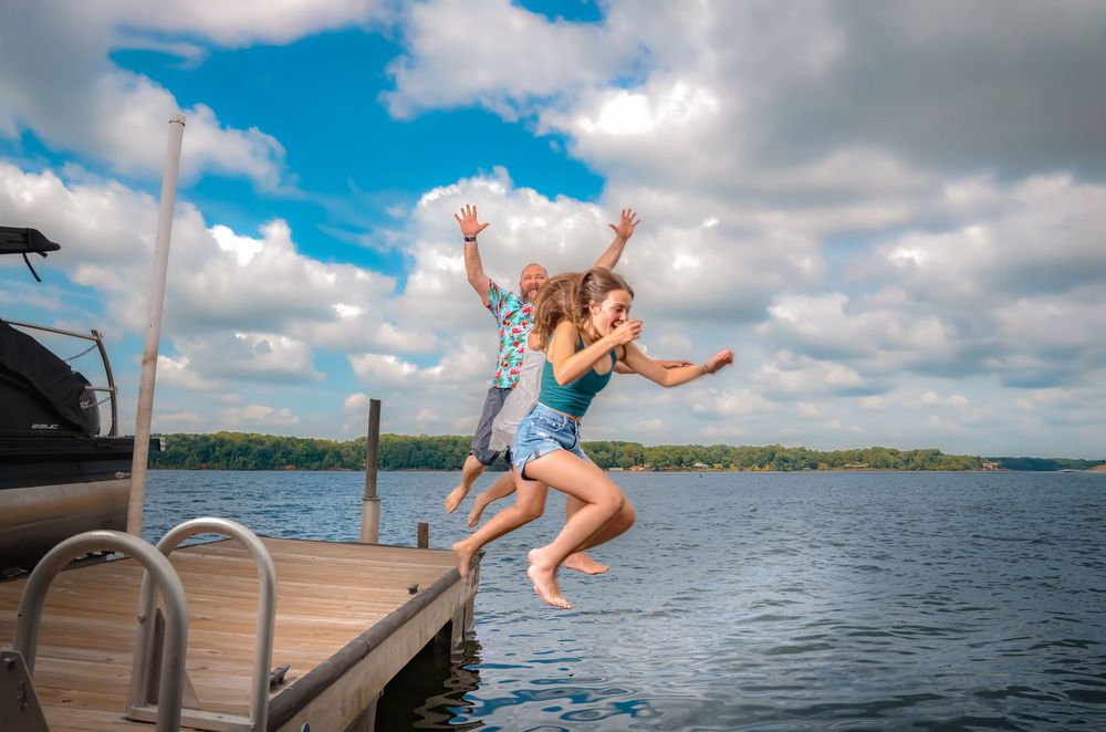 Person in denim shorts joyfully jumps off wooden dock into lake on sunny summer day with puffy clouds overhead.