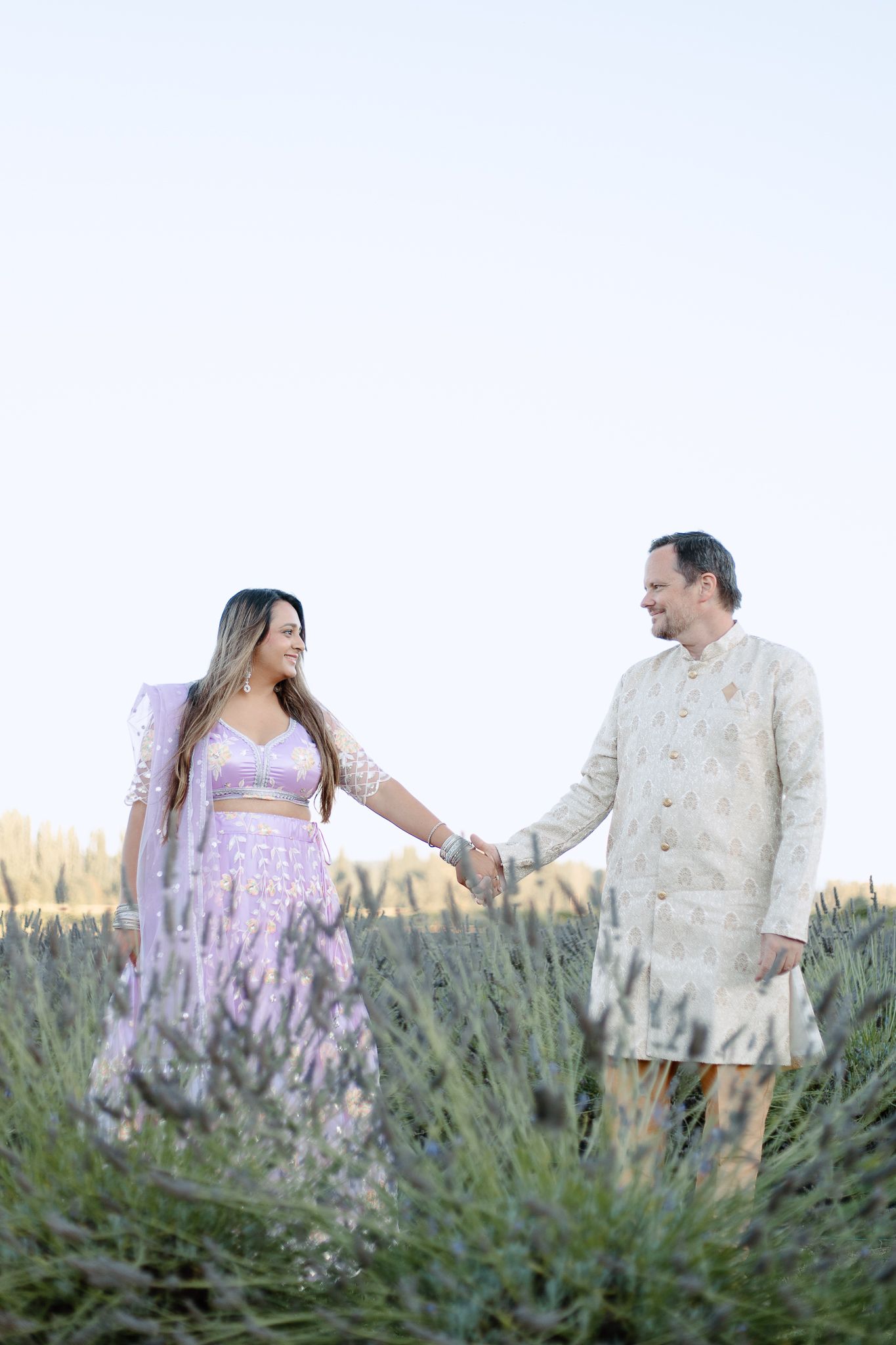 A couple poses together in a lavender field at sunset wearing coordinating white and purple outfits.