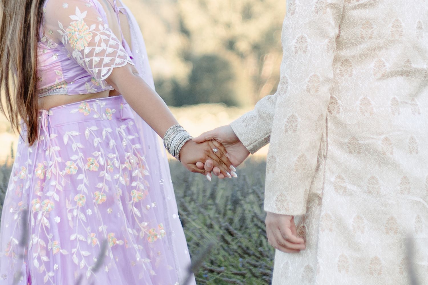 A couple in lavender and white outfits embrace in a romantic outdoor photoshoot among greenery and sunlight.