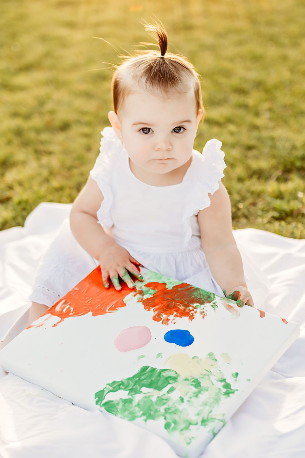 A small child in a white dress sits on a blanket doing colorful finger painting artwork outdoors on a sunny day in Colleyville, TX
