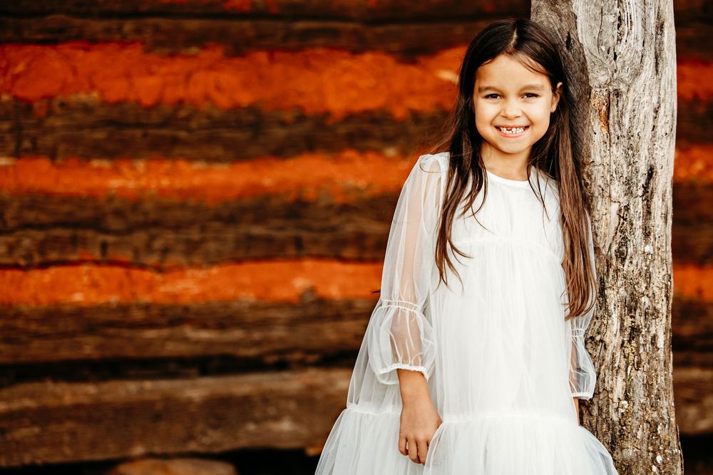 A child in a white dress stands against a rustic log cabin wall with warm autumn lighting in Colleyville, TX