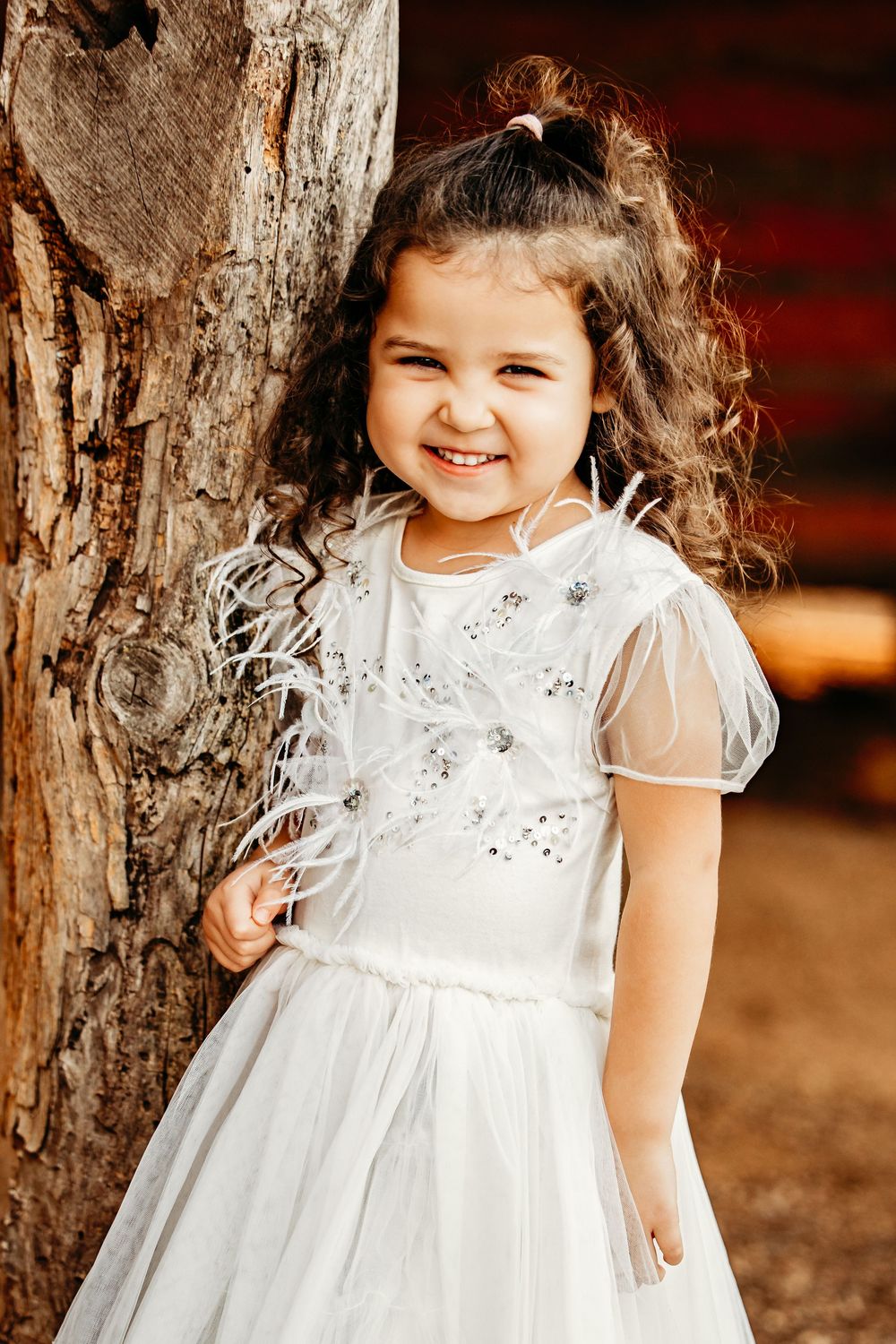 A toddler in a white lace dress poses against a rustic cabin in Colleyville, Texas