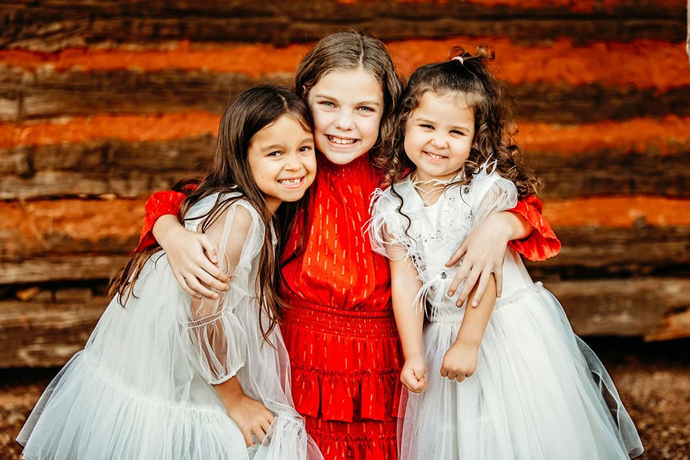 Three children in formal dresses pose together against a rustic wooden wall with warm autumn lighting in Colleyville, TX