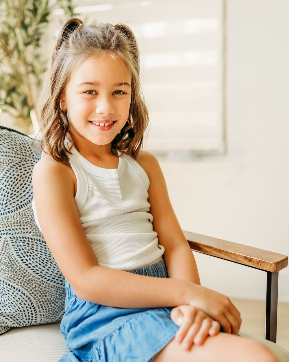 Girl in white tank top and blue skirt sits on chair with decorative pillows in bright interior setting for an in-home session in DFW