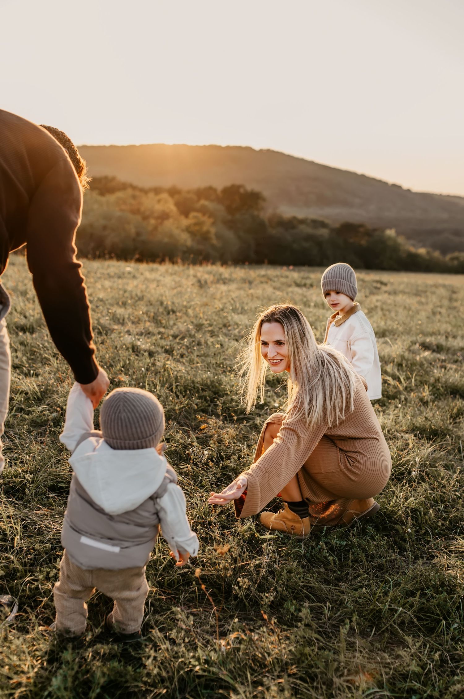 A toddler in a knit beanie explores a grassy hillside at sunset while someone holds their hand for support.