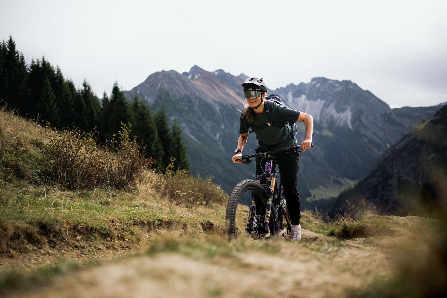 Mountain bikers traverse scenic alpine trails with rugged peaks and dramatic mountain landscapes in the background.