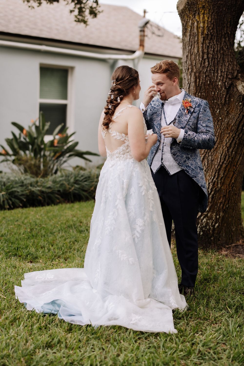A couple shares a romantic moment in their wedding attire under a tree on a grassy lawn.
