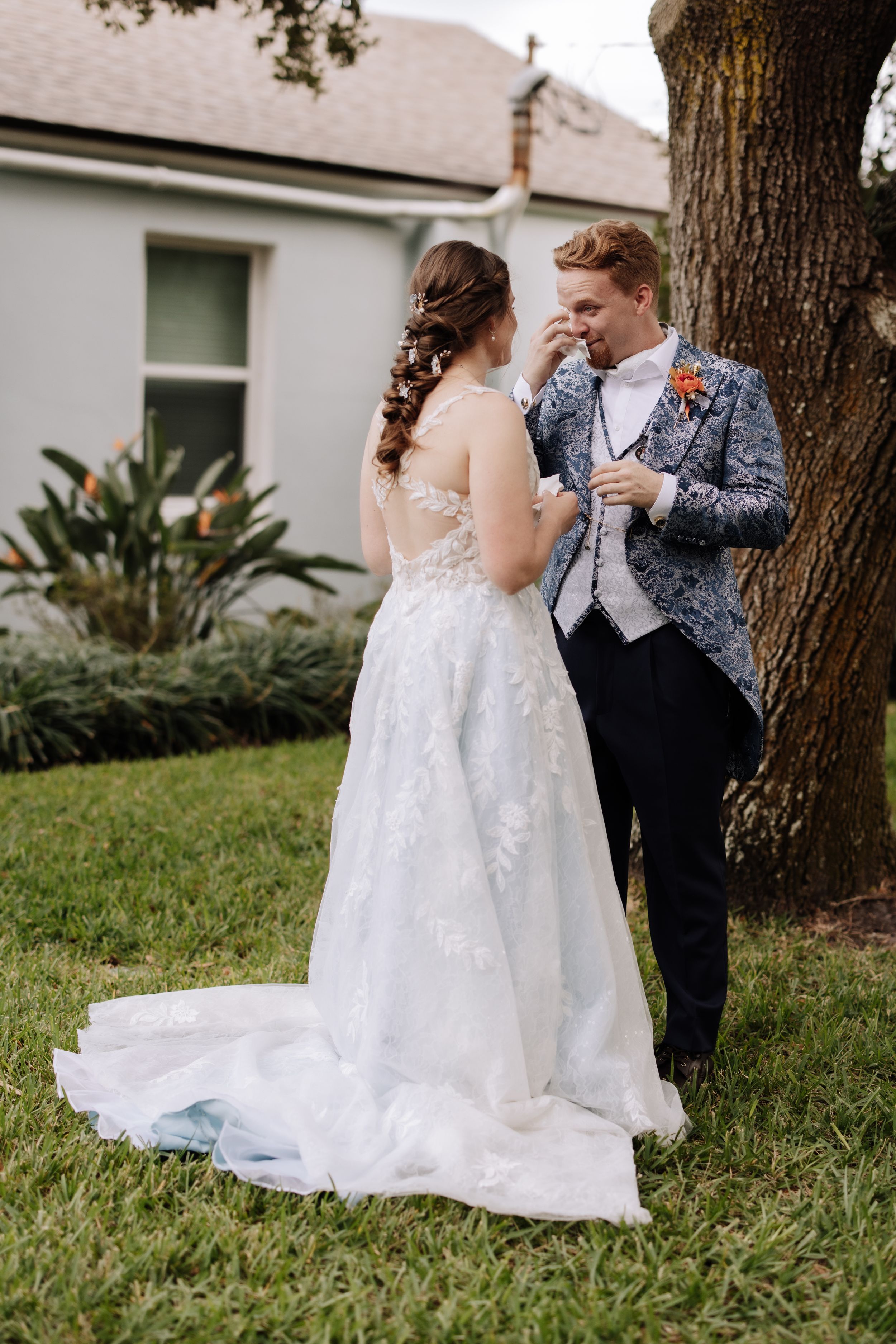 A couple shares a romantic moment in their wedding attire under a tree on a grassy lawn.