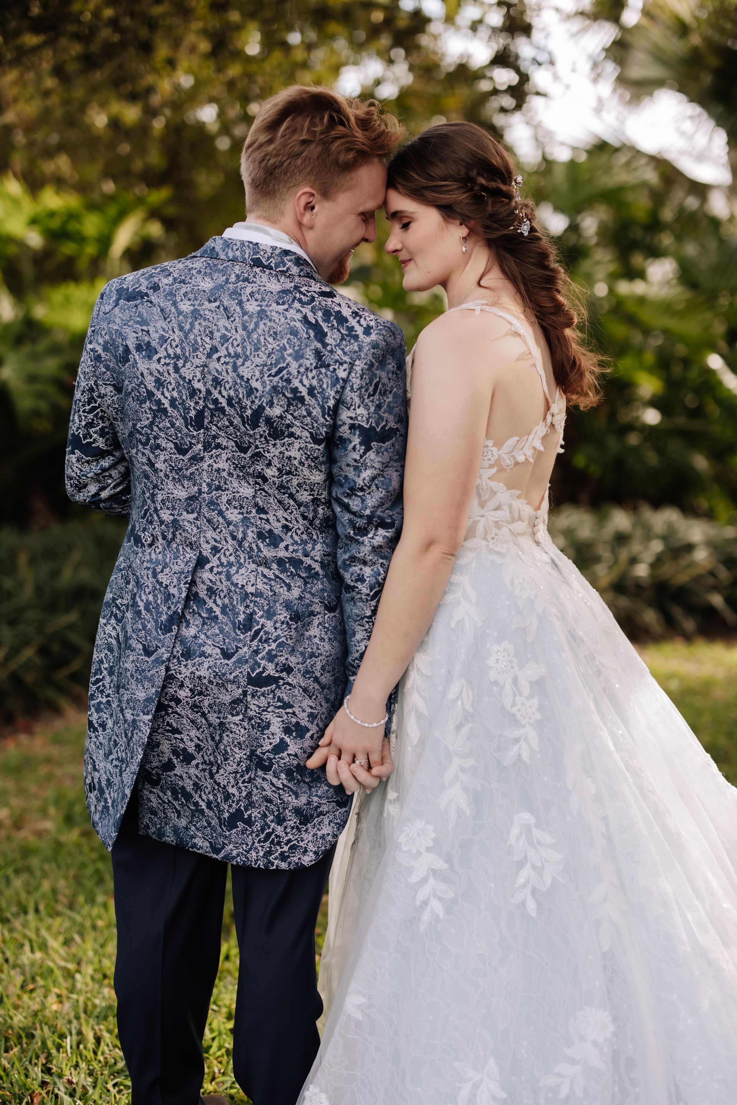 A romantic wedding couple shares an intimate moment in a garden wearing a blue floral suit jacket and lace gown.