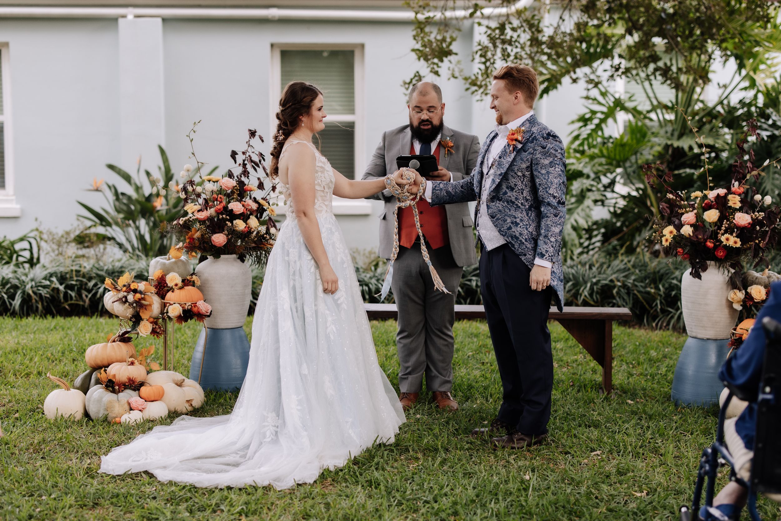 Outdoor wedding ceremony with floral decorations and guests gathered on a lawn in front of a white building.