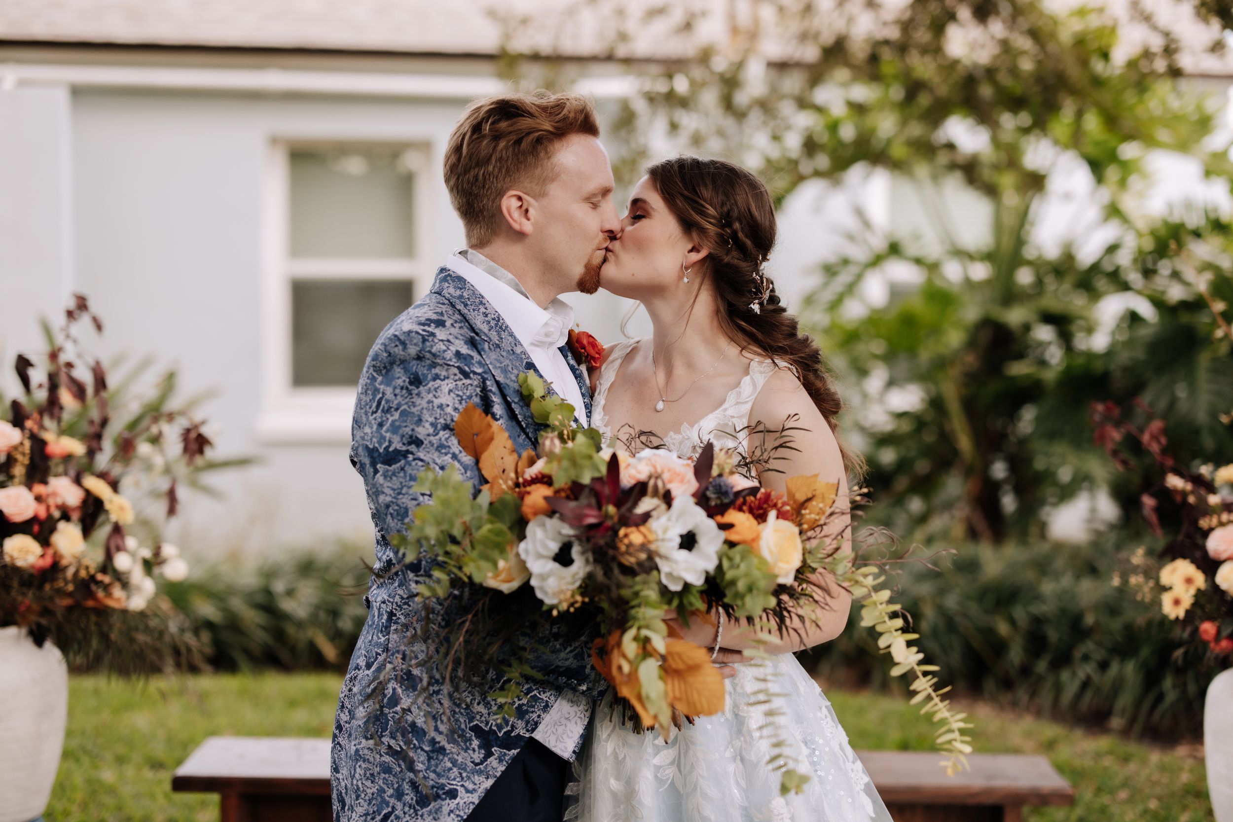 A romantic wedding couple shares a tender kiss in a garden setting surrounded by autumn flowers and greenery.