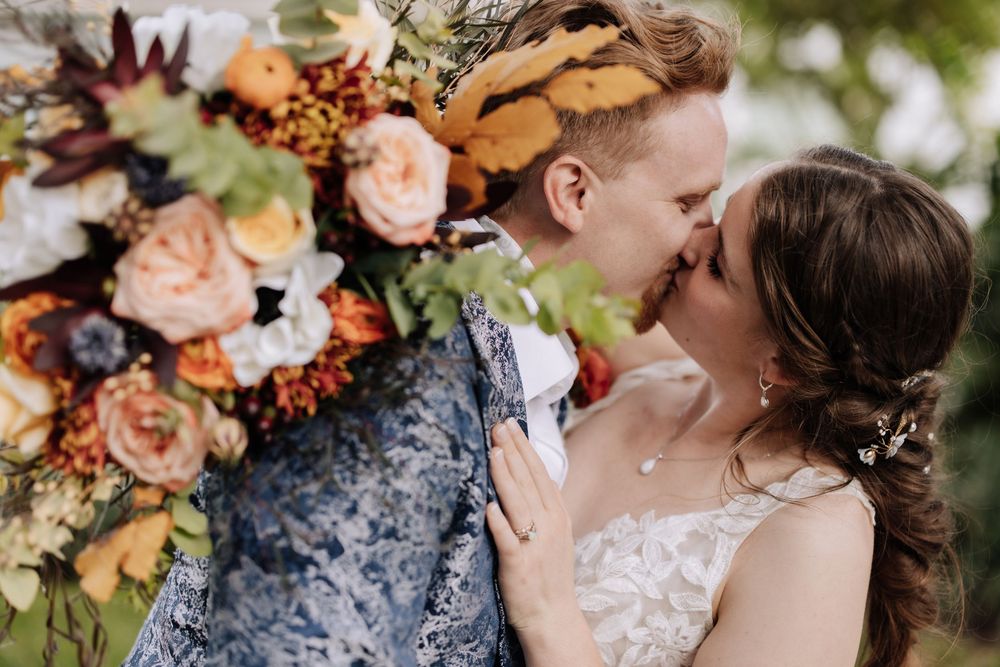 A romantic wedding couple shares an intimate kiss surrounded by peach and coral floral arrangements in a garden setting.