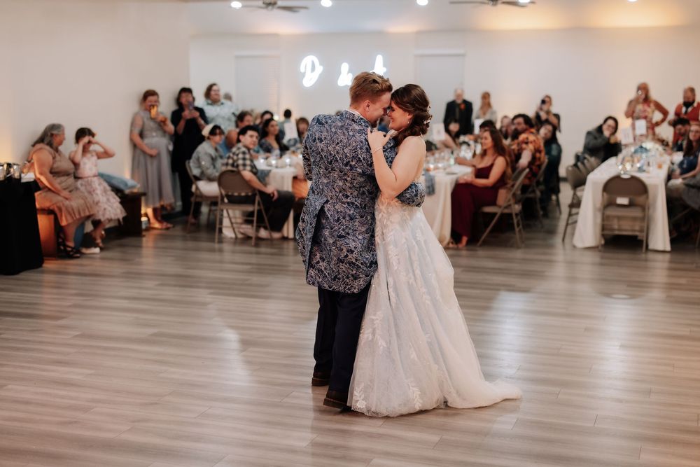 A couple shares a romantic dance together on a wooden floor at a wedding reception as guests look on from their tables.