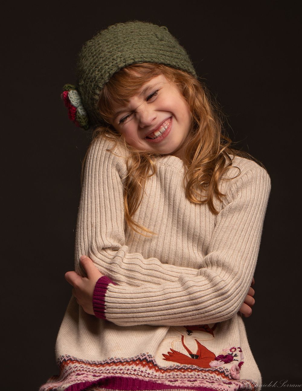 A cheerful young girl in green knit hat and sweater hugs herself smiling warmly.