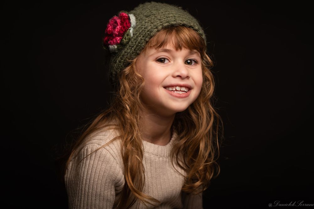 A smiling young girl with long hair wearing a green knit hat with flower detail.