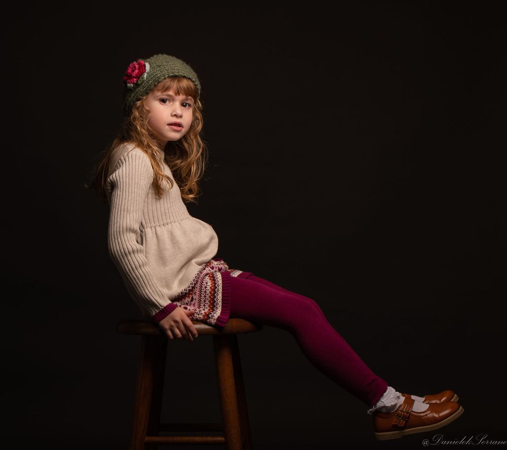 Young girl in knit hat and sweater sitting on wooden stool against dark background.