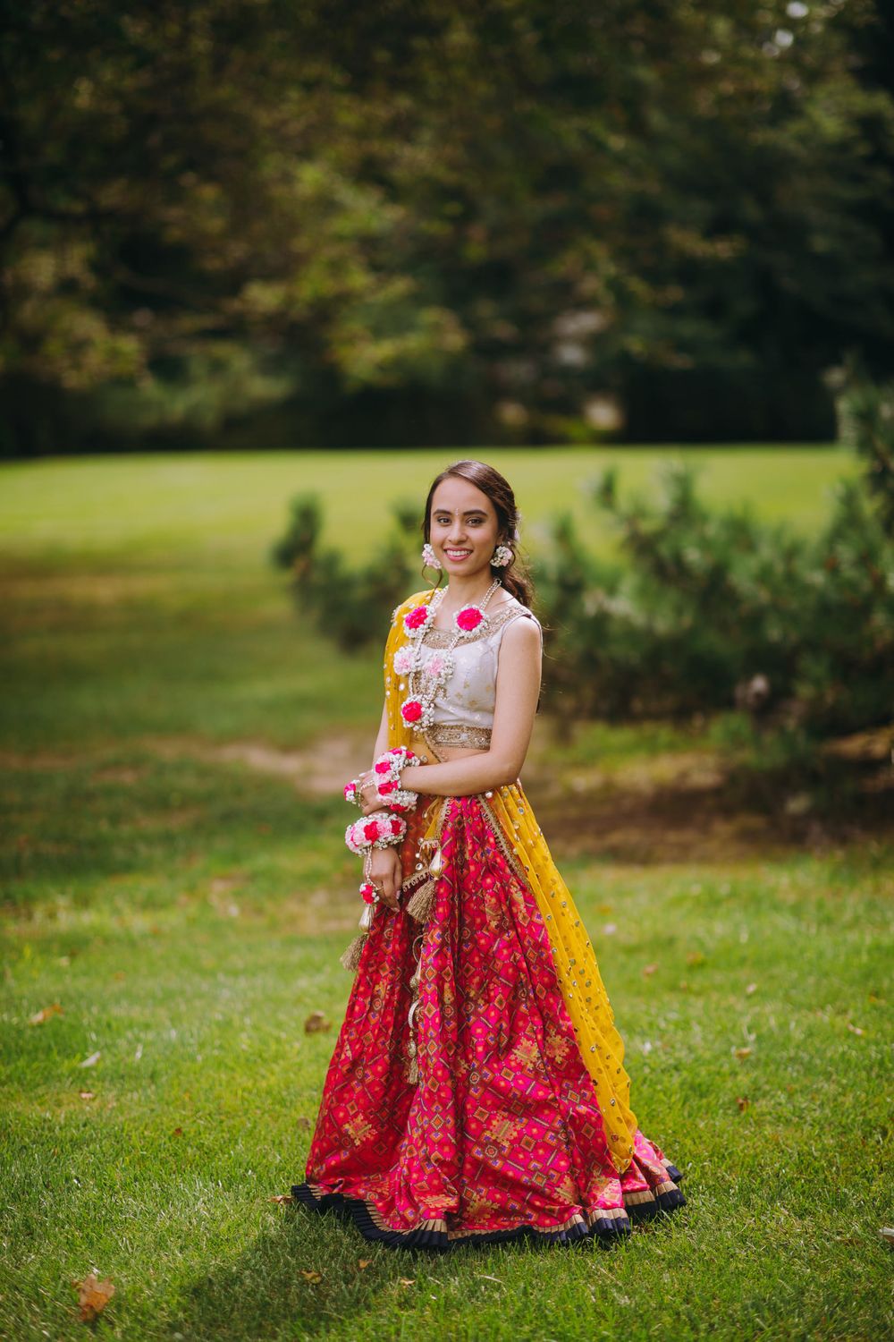 A person in a vibrant red and yellow traditional Indian lehenga poses gracefully on a lush green lawn.
