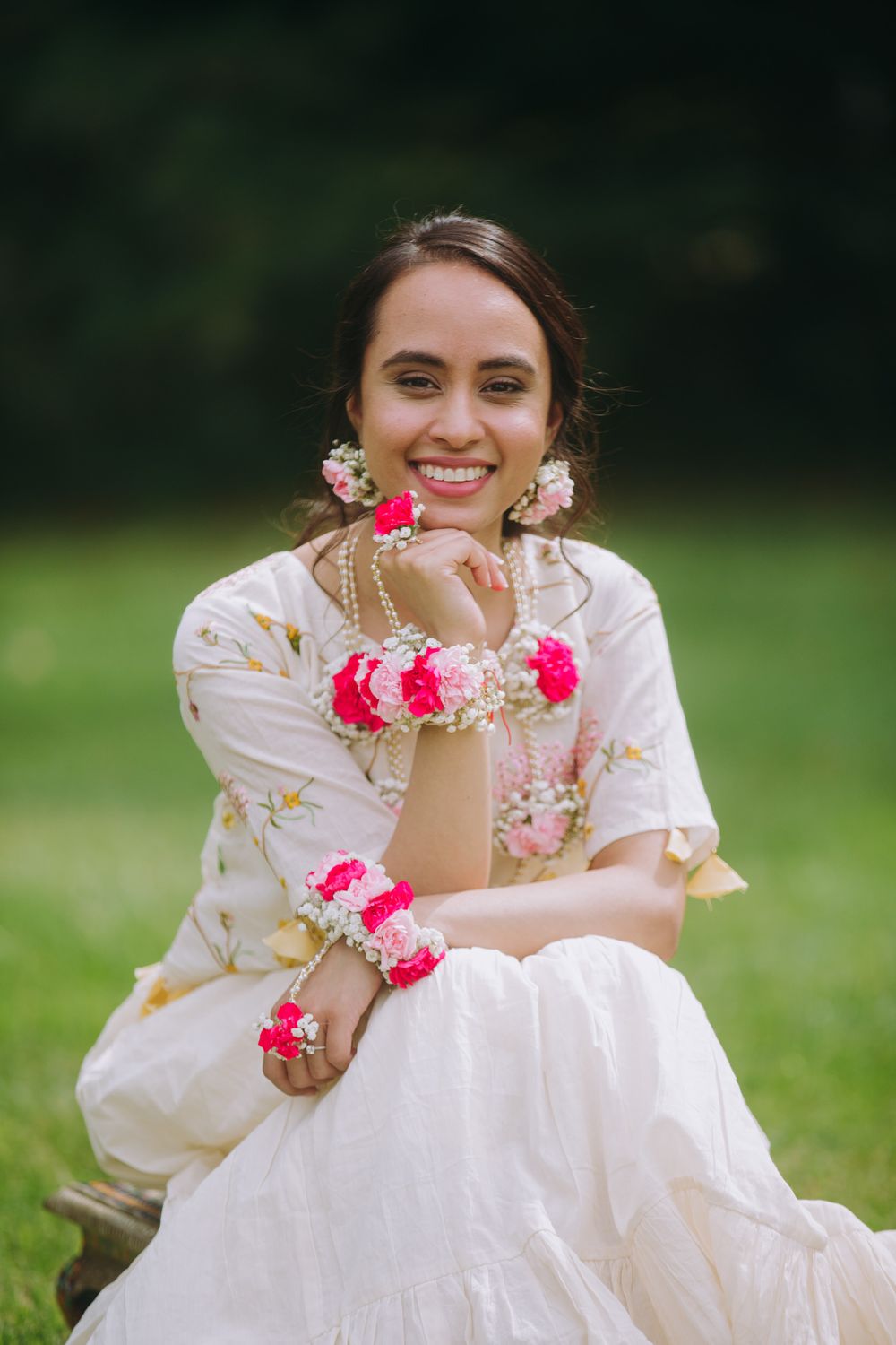 A person in a white dress with pink floral accessories poses joyfully on a grassy lawn.