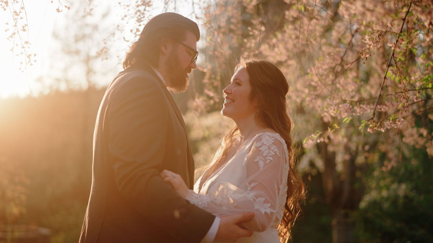 A romantic couple embraces outdoors during golden hour sunlight with cherry blossoms blooming in the background.