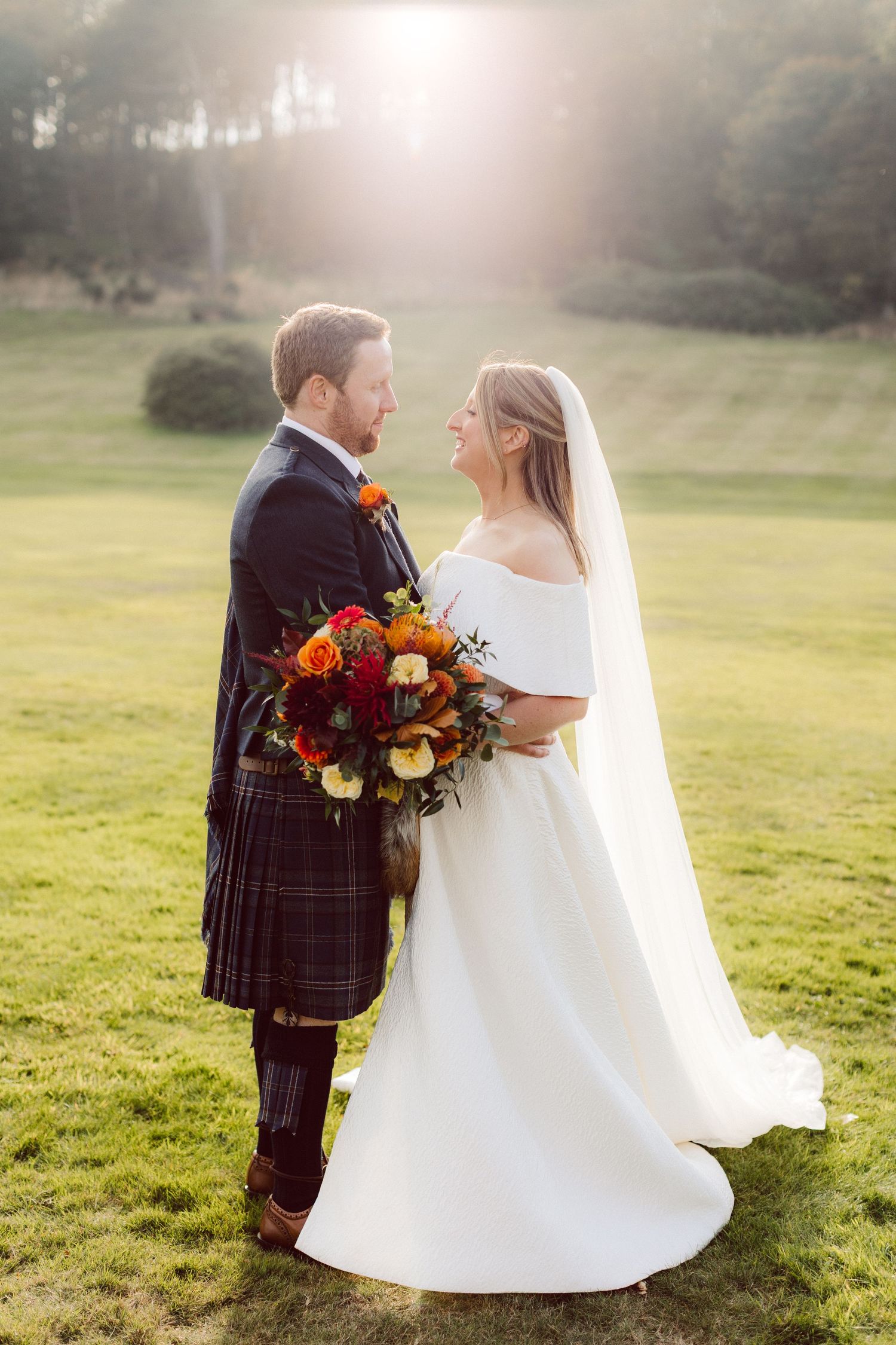 Sunlit wedding portraits with bride in white dress and groom in traditional Scottish kilt.