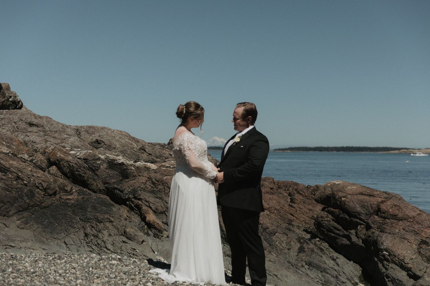 Bride and groom hold hands facing each other during ceremony on a rocky coastal beach with ocean backdrop.
