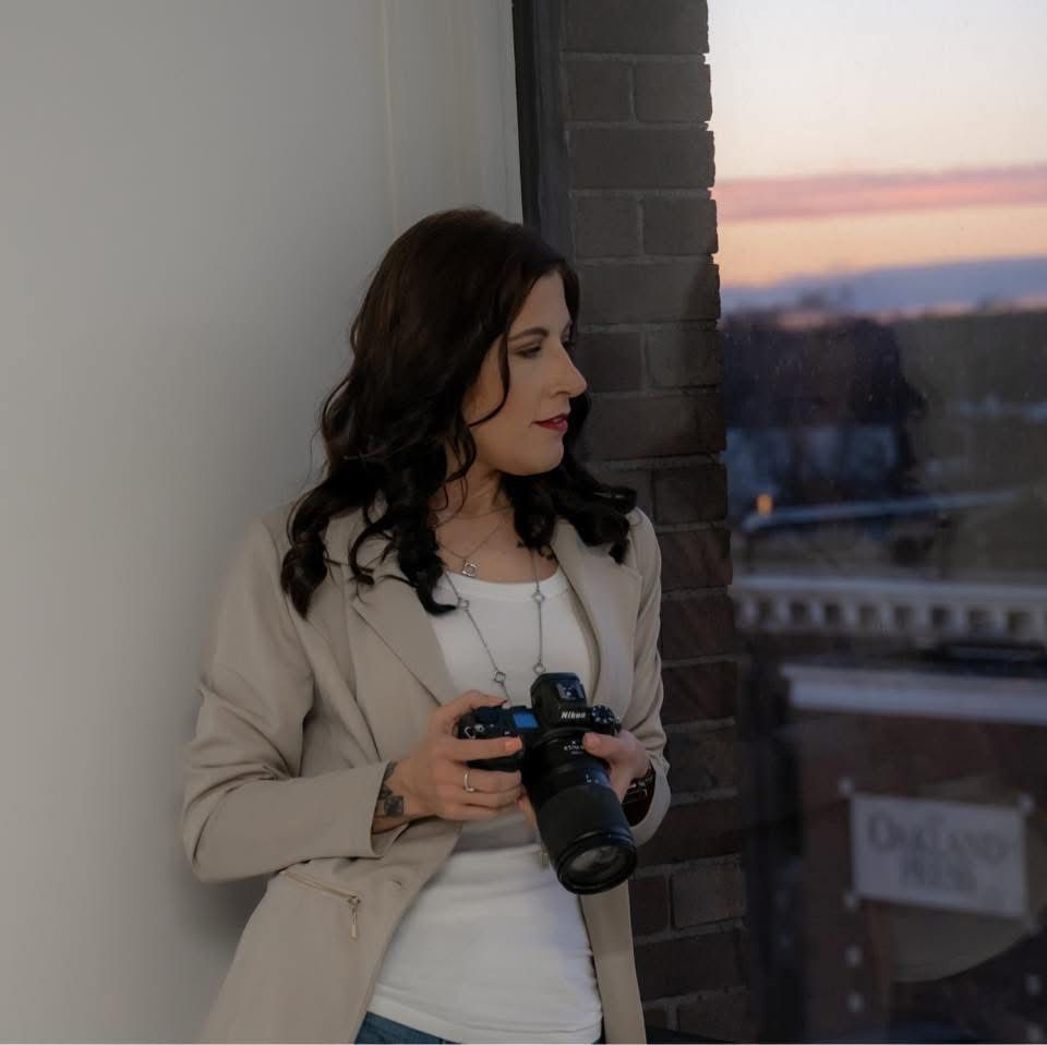 Woman photographer holding camera near brick wall at sunset, wearing beige blazer and white top.
