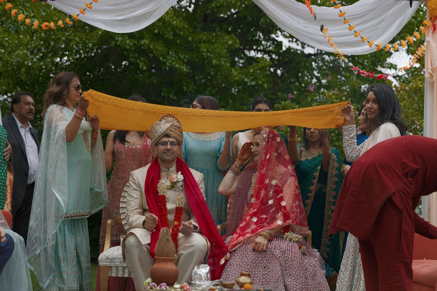 Bride and groom seated under colorful mandap canopy during traditional outdoor Indian wedding ceremony.