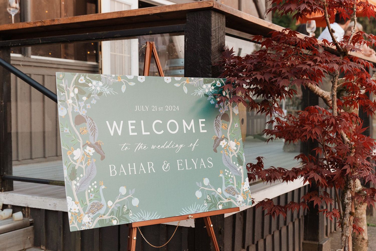 Wedding welcome sign for Bahar and Elyas displayed on easel beside red Japanese maple tree.