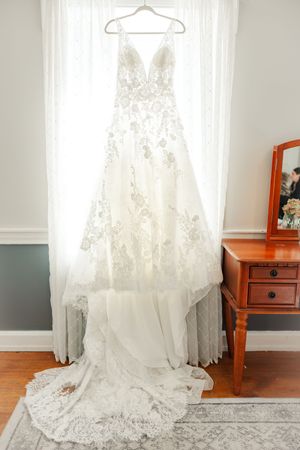 Detail, Wide, Color, Neutral, 2. Elegant lace wedding dress displayed on hanger against white curtains in sunlit room.