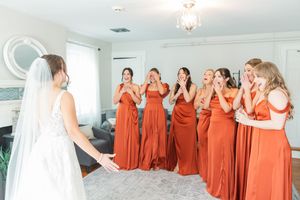 Bridesmaids in matching burnt orange dresses share emotional moments with the bride in a bright indoor setting.