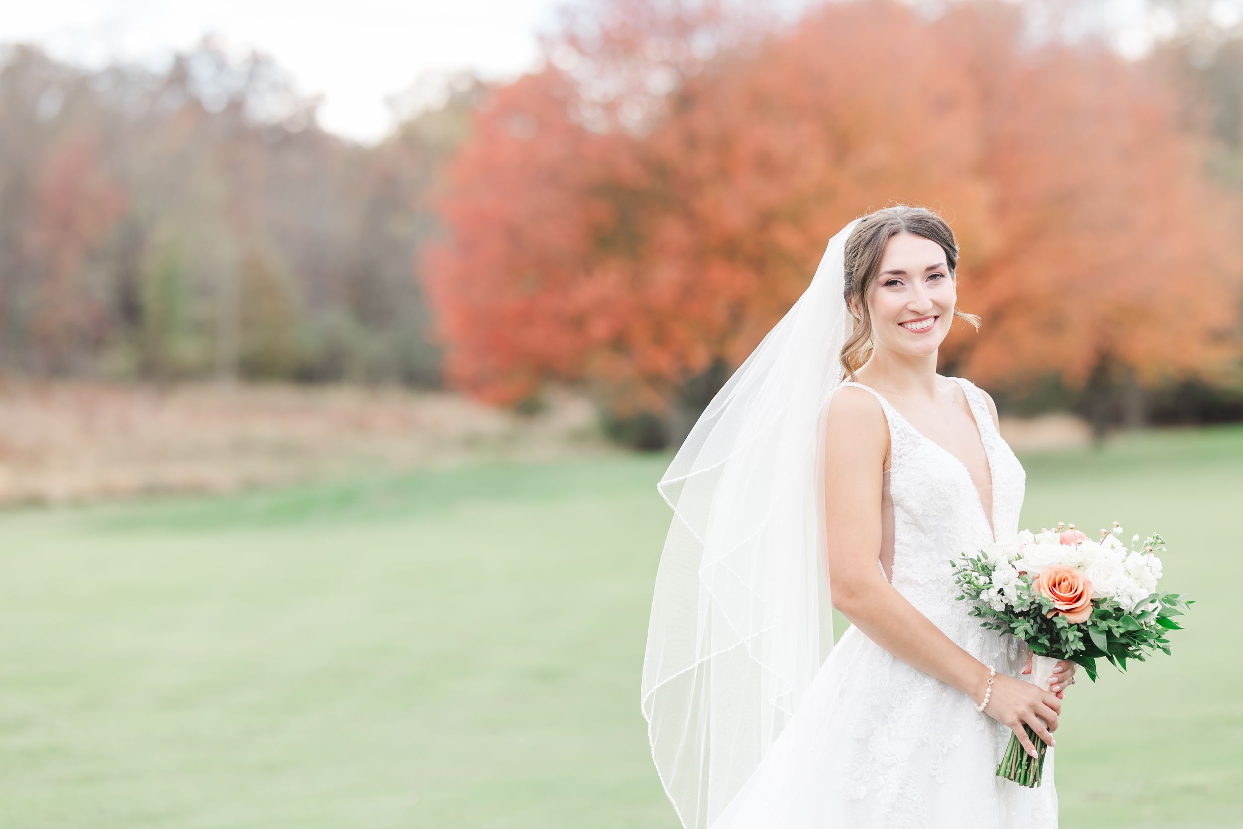 Bride in white dress holding bouquet against autumn foliage background.