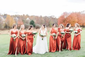 Bride in white dress with bridesmaids wearing rust-colored dresses pose in fall landscape.