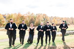 Groomsmen in black tuxedos pose in a fun line formation on green grass during fall season.