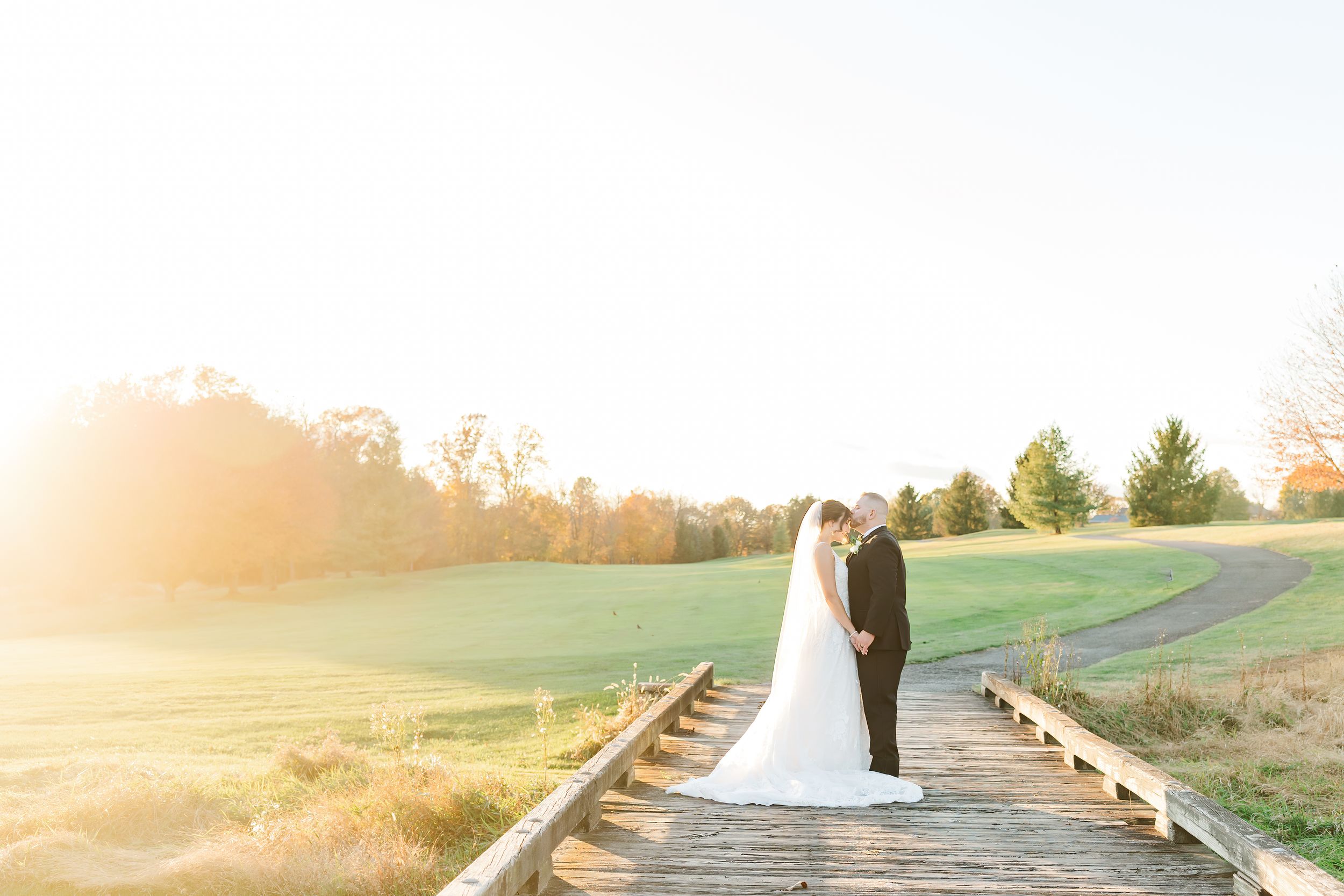 Bride and groom walk along a sunlit path through rolling green hills during golden hour sunset.