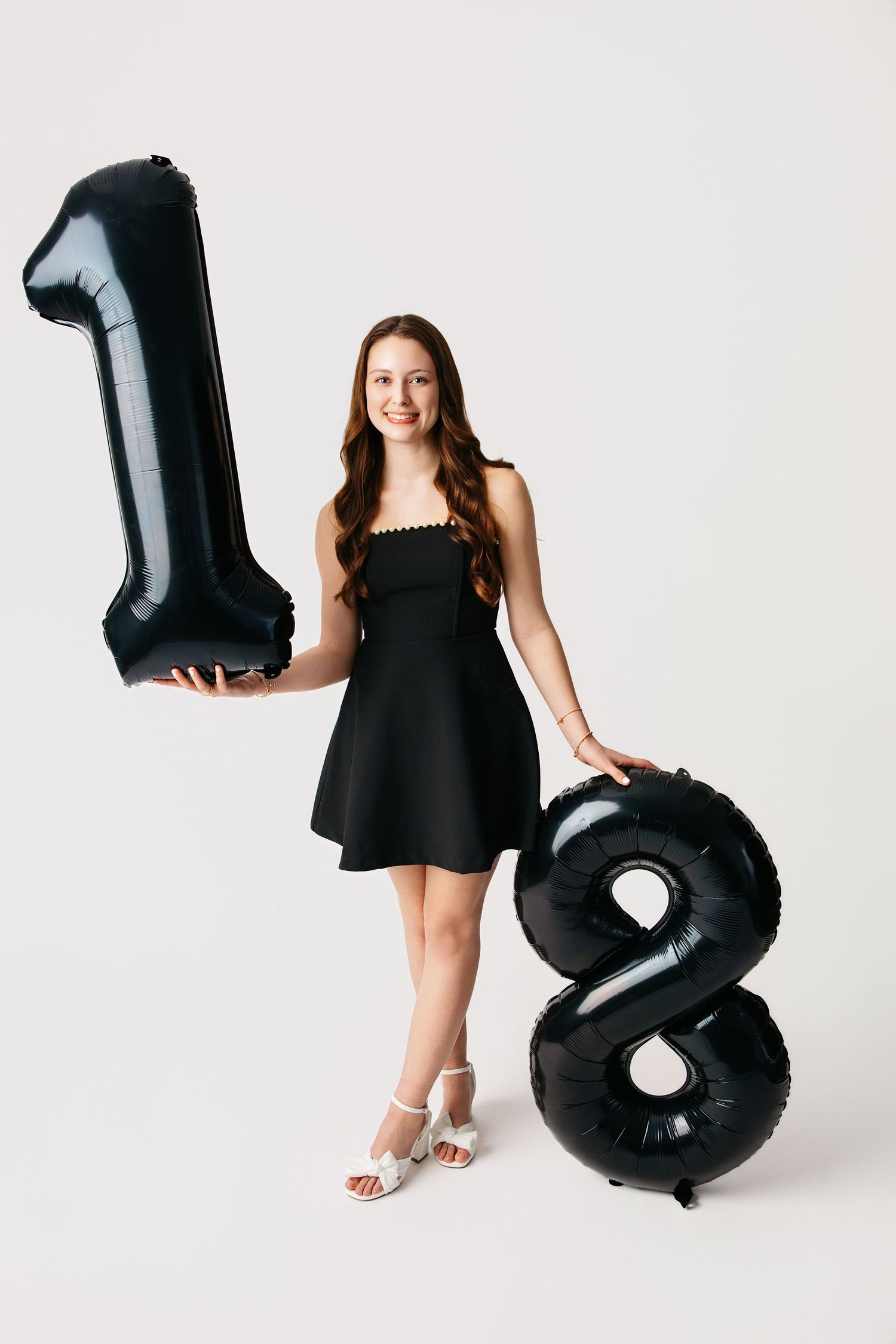Young woman in black dress poses with large black number balloons showing 18 against white background.