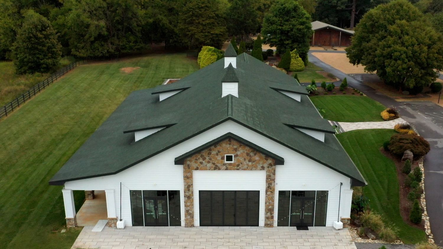 Aerial view of a green-roofed ranch style home with attached garage and circular driveway surrounded by landscaping.
