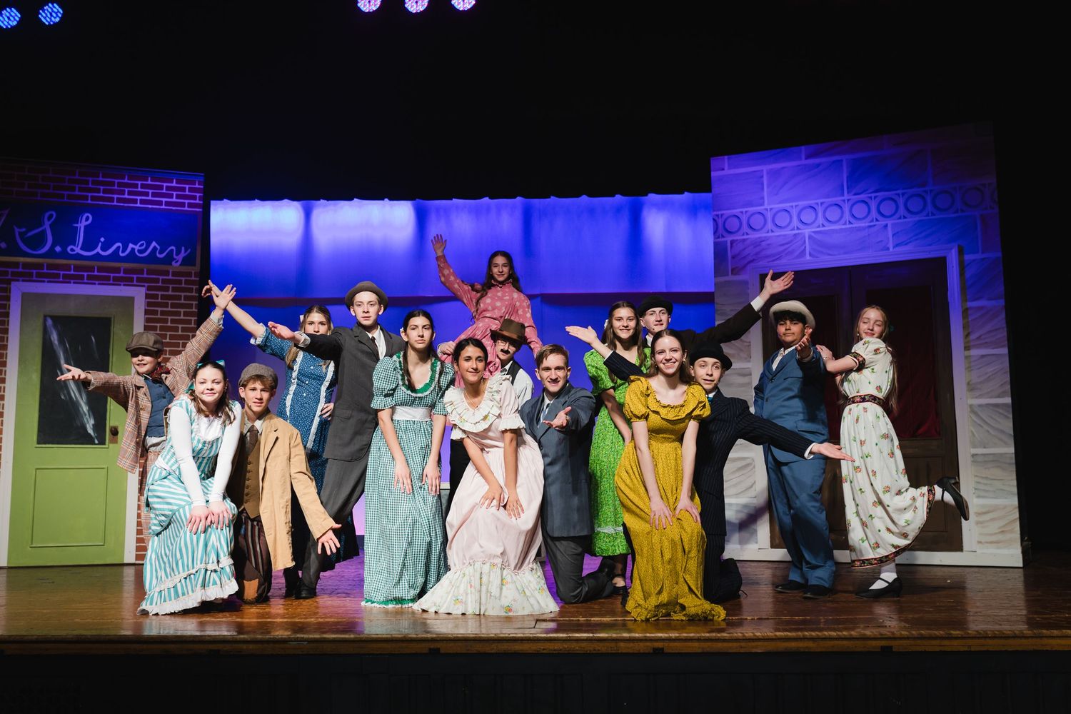 Cast members perform on stage in period costumes during a theatrical production with blue lighting effects.