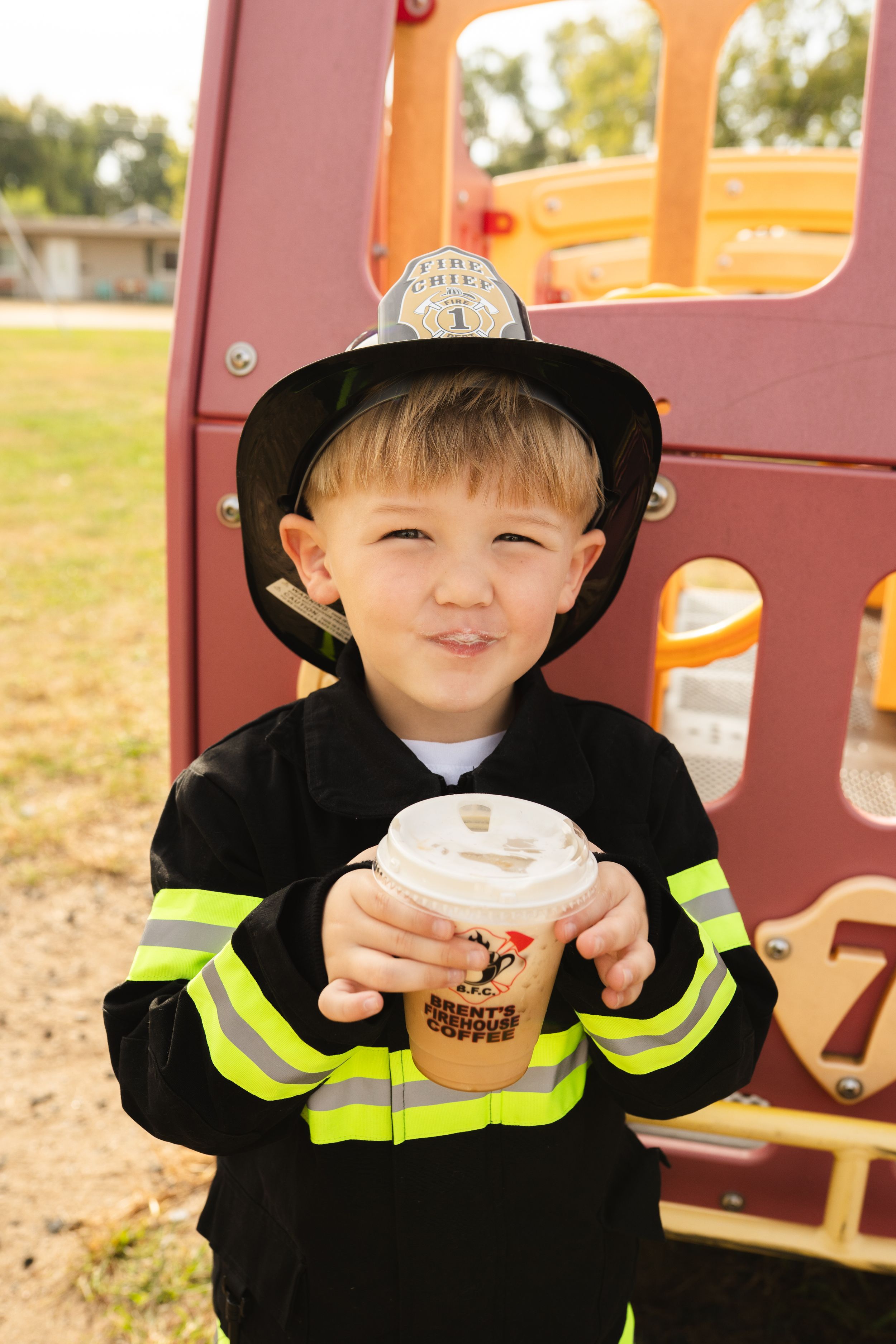 Figure in firefighter costume holding a Dunkin' Donuts cup near playground equipment.