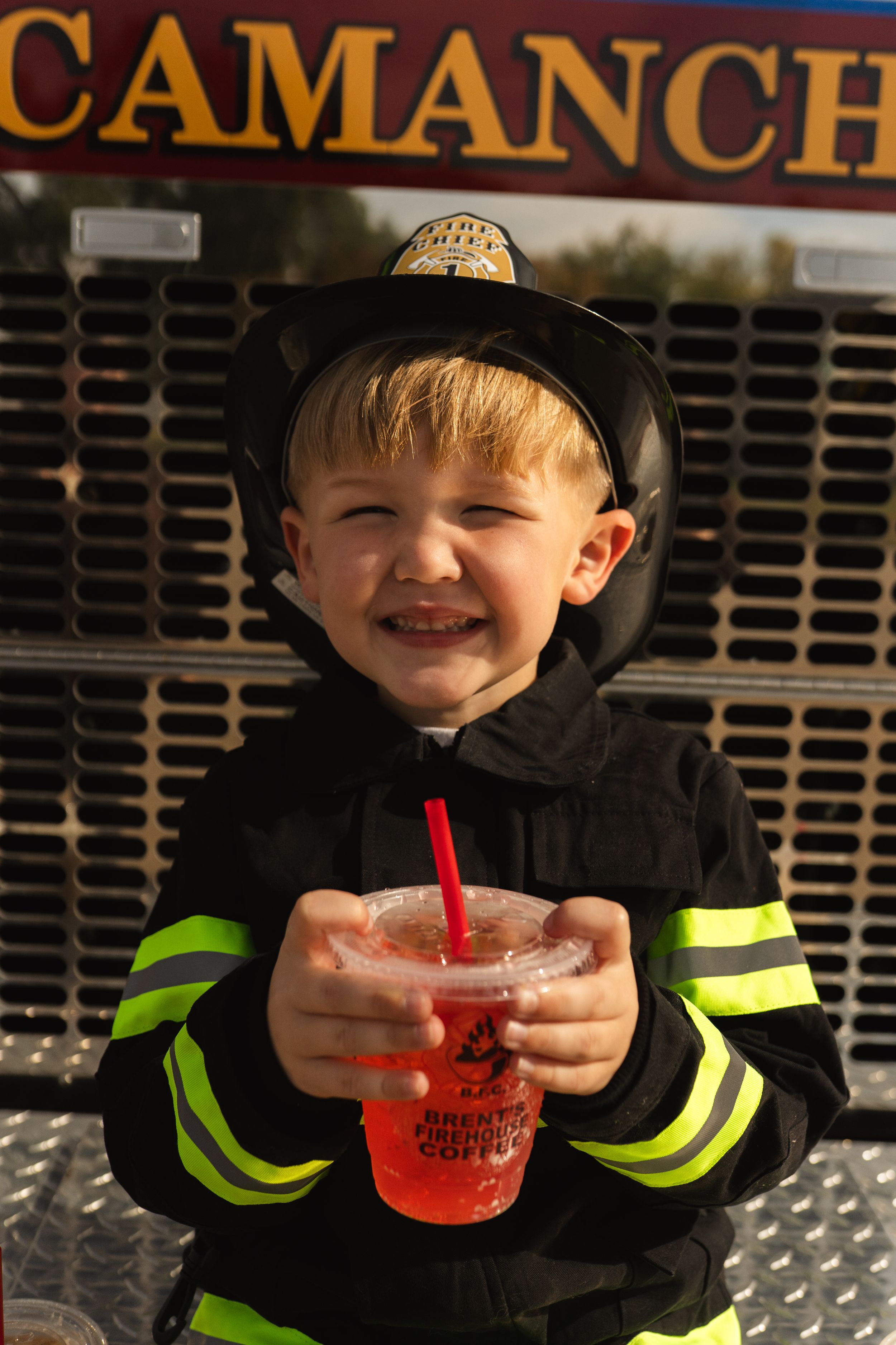 Figure in firefighter costume drinking red beverage through straw near Camanch sign.