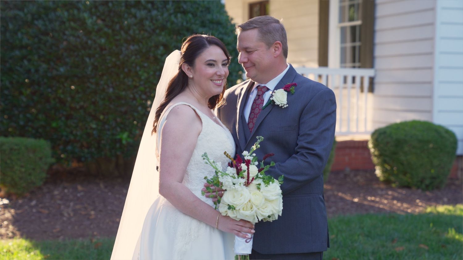 Bride and groom share intimate moments in outdoor garden setting with white church building in background.