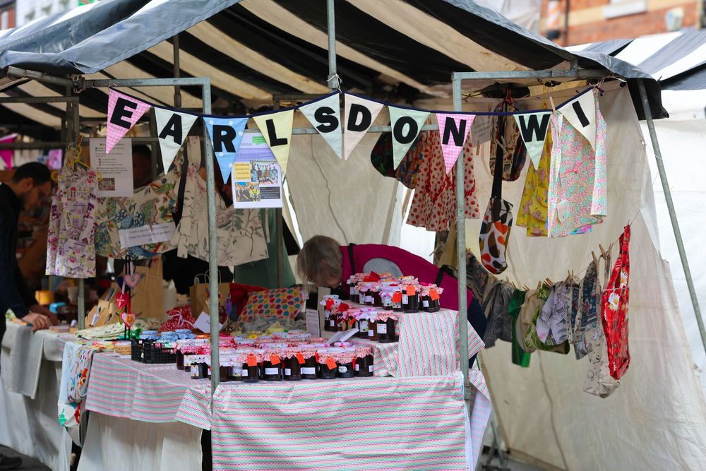 Earlsdon market stall with colorful bunting, handmade clothing, and jars displayed under a striped canopy tent.