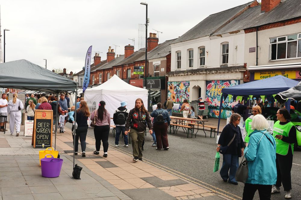 Busy outdoor street market with vendor tents, shoppers browsing stalls, and colorful storefronts on a cloudy day.