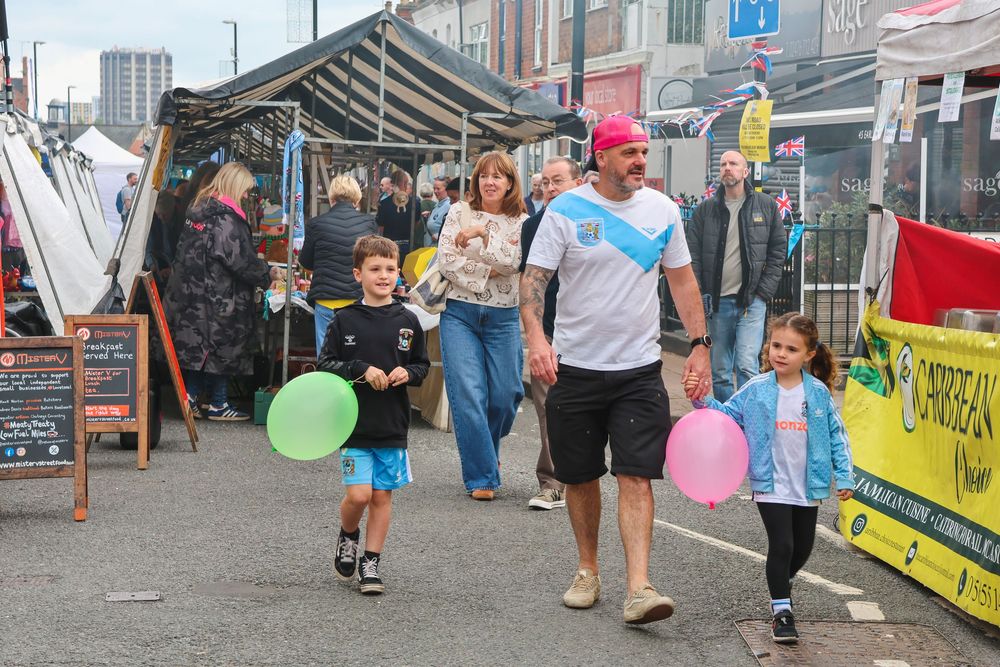 Family with balloons strolling through a vibrant outdoor street market with stalls and bunting on a cloudy day.