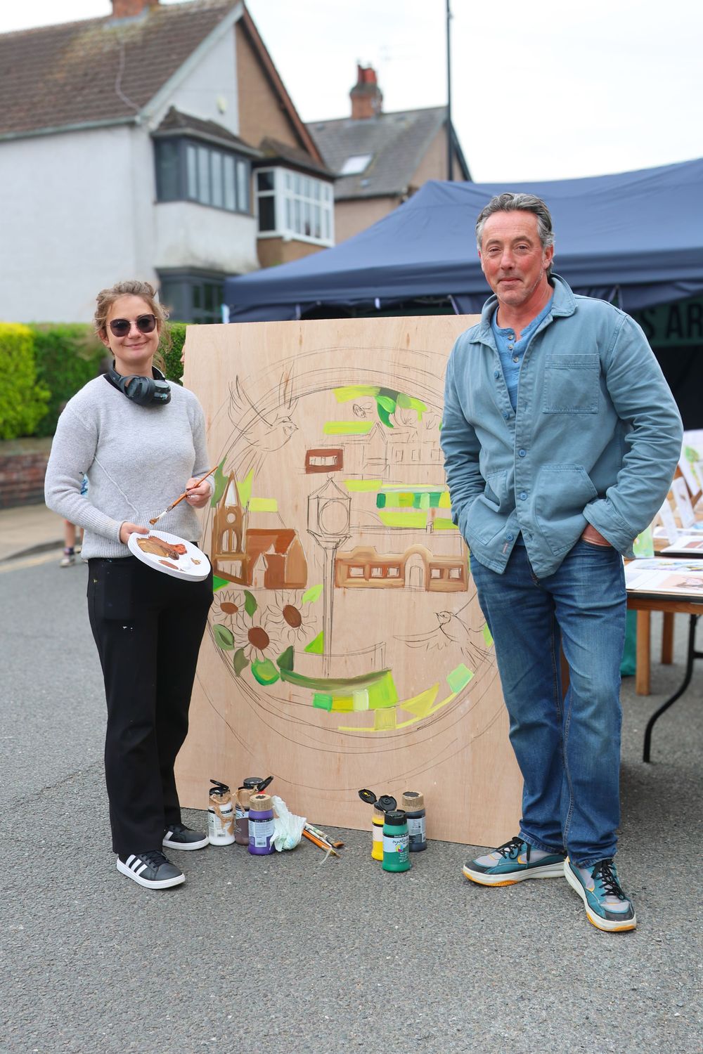 Two people posing beside a large colorful painted mural artwork at an outdoor community festival event.