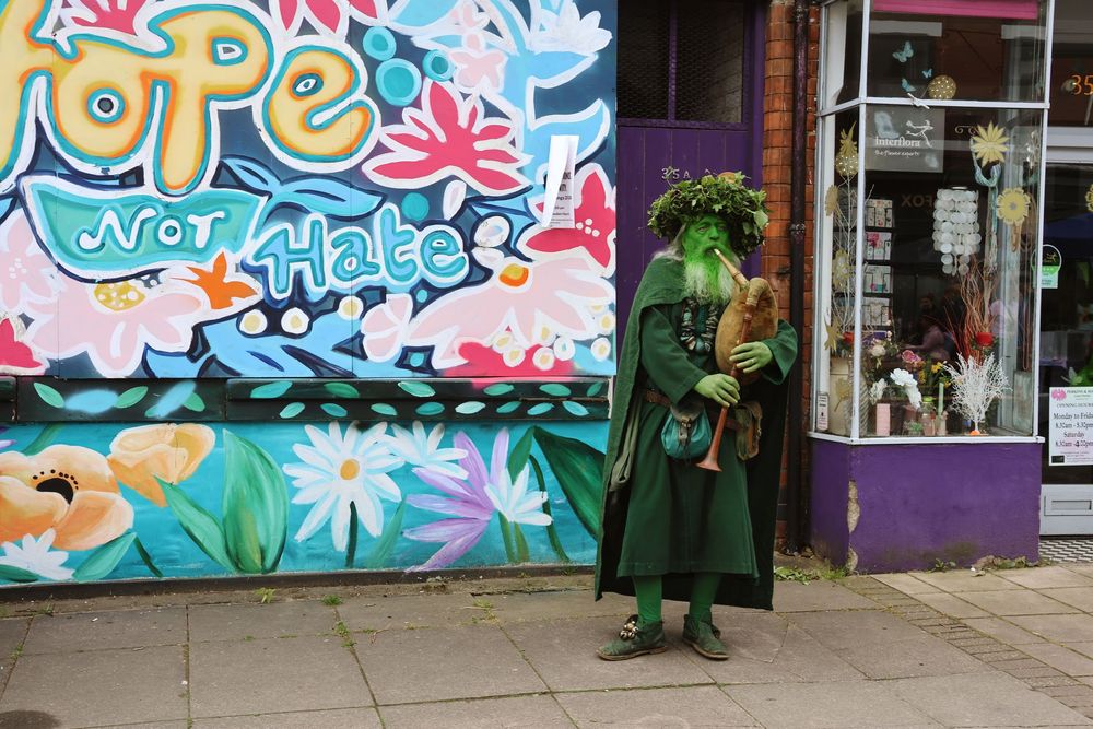 Person in green costume standing before a Hope Not Hate mural with colorful flowers on a shop wall.