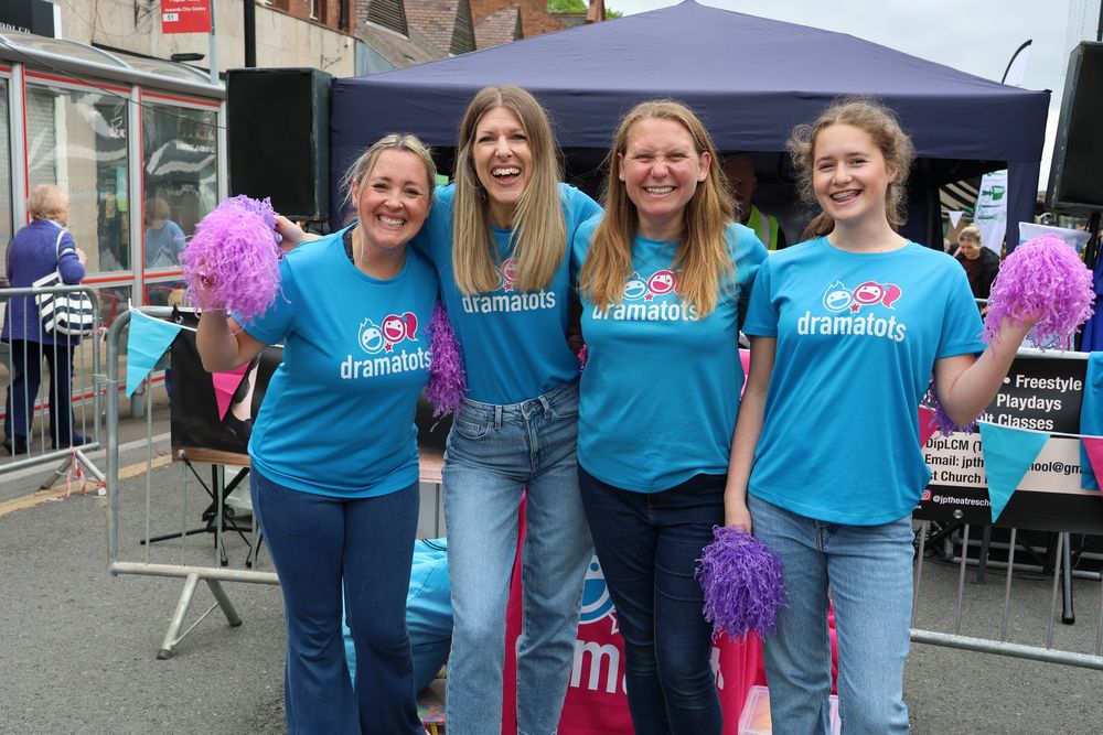 Four women in blue Dramatots t-shirts smiling and holding purple pom-poms at an outdoor community event.