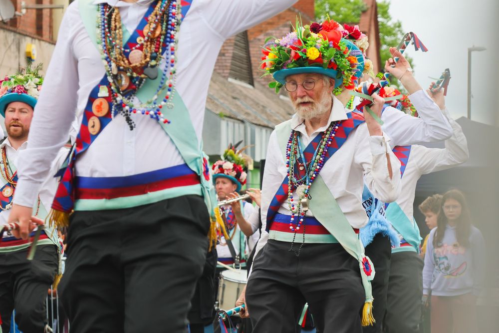 Morris dancers in colorful flower crowns and traditional costumes performing at an outdoor street festival.
